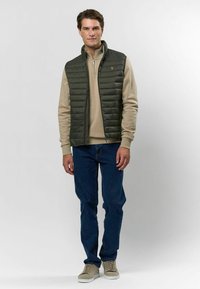 Olivnozeleni prešani prsluk preko bež dukserice, upareni s plavim farmerkama i sivim visokim tenisicama. Detalj s patentnim zatvaračem na ovratniku prsluka.