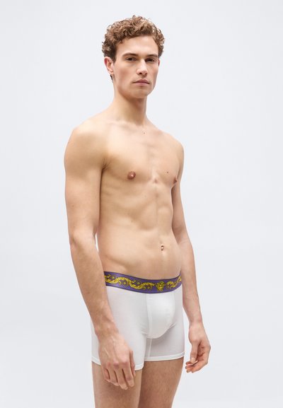 Νέος άνδρας με σγουρά μαλλιά, όρθιος χωρίς πουκάμισο, φοράει λευκά μπόξερ με μωβ και χρυσή ζώνη με σχέδια.