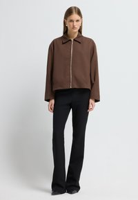 Brun cropped jakke med lynlås foran og spredt krave, parret med sorte tætsiddende bukser, begge i glatte stoffer.