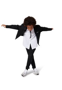 Giacca nera con ampi revers, camicia bianca e pantaloni neri. Sneakers grigie e bianche notevoli, tessuto texturizzato e design minimalista.