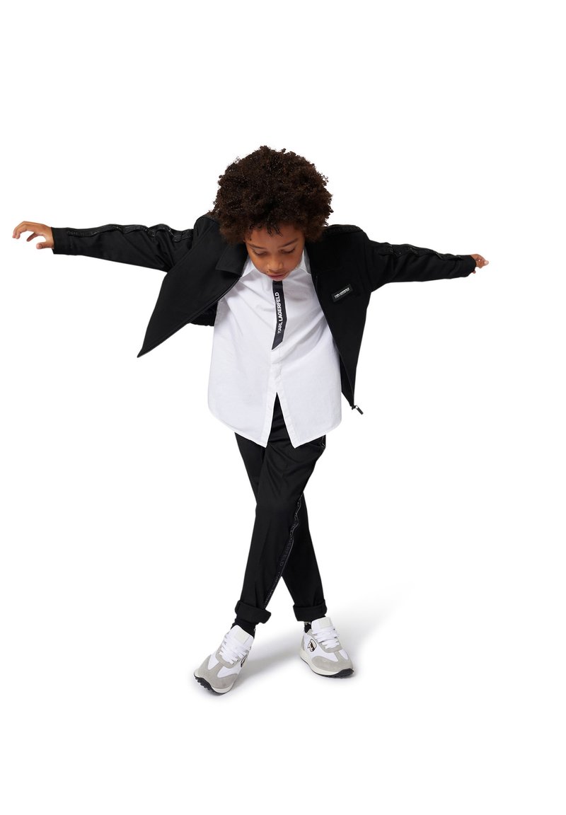 Giacca nera con ampi revers, camicia bianca e pantaloni neri. Sneakers grigie e bianche notevoli, tessuto texturizzato e design minimalista.