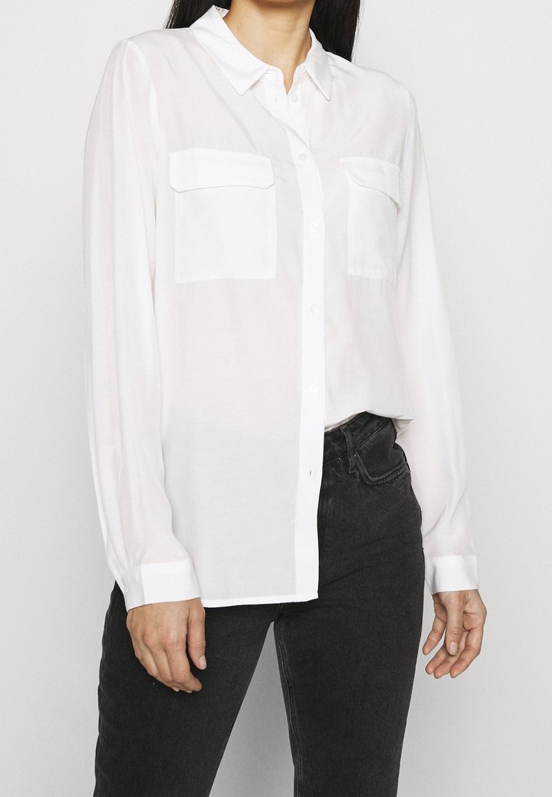 Chemise blanche à manches longues boutonnée avec deux poches poitrine portée avec un jean noir, montrée de l’épaule à la hanche sur une personne.