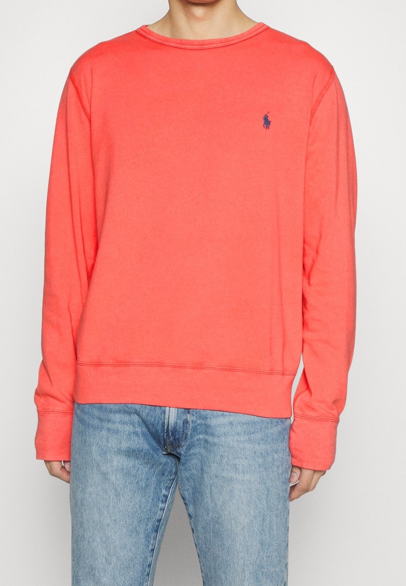 Homme portant une chemise corail à manches longues avec un petit logo polo bleu marine sur la poitrine et un jean bleu clair, debout devant un fond uni.