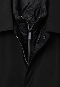 Veste noire pour homme avec une fermeture éclair partiellement ouverte à l'avant et une couche intérieure rembourrée, montrant une étiquette avec la mention "Calliope Man L."