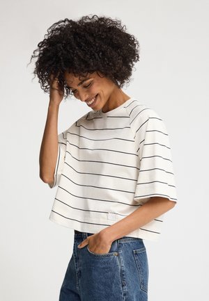 Kvinde smilende med krøllet hår, iført en hvid kortærmet skjorte med tynde sorte vandrette striber og blå jeans, hånd i lommen.