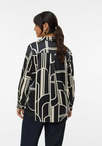 Black and cream long-sleeve blouse featuring a geometric pattern, soft fabric, and a relaxed fit; detailed button cuffs and back pleat.