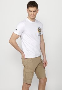 White cotton t-shirt with short sleeves features a graphic print in black and orange. Paired with beige cargo shorts with multiple pockets.