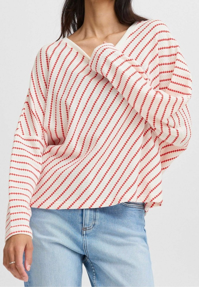 Pull-over décontracté blanc avec des rayures diagonales rouges en V. Texture douce, manches longues et poignets côtelés. Porté avec un jean bleu clair.