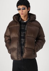 Chaqueta de plumón marrón con capucha, con diseño acolchado, textura brillante y cierre de cremallera, llevada sobre una camisa a cuadros en negro y gris.
