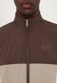 Hnědá a béžová bunda na zip s vysokým límcem, která má texturovanou látku a logo EA7 na levé straně. Zip je viditelný.