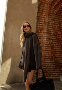 Übergröße, dunkelgrauer Strickpullover mit Kapuze, langen Ärmeln und lockerer Passform. Kombiniert mit dunklen Shorts und einer strukturierten schwarzen Tragetasche.