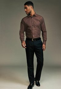 Cămașă cu mânecă lungă, maro cu model, pantaloni negri și mocasini negri cu accente aurii; poziție în picioare pe un fundal neutru.