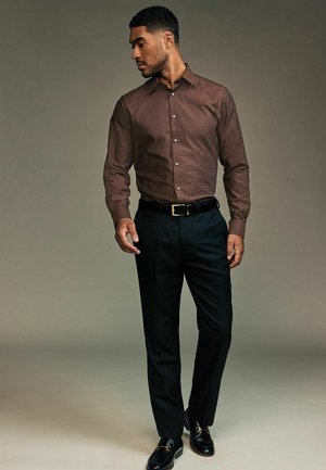 Braun gemustertes Langarmhemd, schwarze Hose und schwarze Slipper mit goldenen Akzenten; stehende Pose vor einem neutralen Hintergrund.