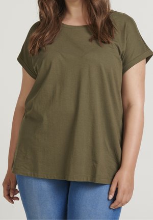 Kobieta ubrana w oliwkowo-zielony krótki rękaw t-shirt i niebieskie dżinsy, stoi na tle jednolitego jasnego tła.