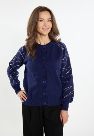 Marineblauwe cardigan met ronde halslijn en knoopsluiting. Kenmerkt zich door textuur en metallic gouden accenten in een swirlpatroon op de mouwen.