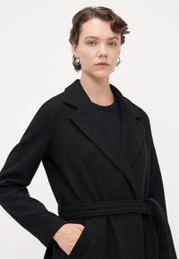 Giovane con capelli corti e ricci, che indossa orecchini a cerchio argento, un cappotto nero con cintura e una maglia nera, posato su uno sfondo bianco.