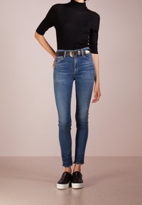 Skinny jeans in medium blue denim with slight distressing at the ankles, paired with a black ribbed turtleneck and black slip-on shoes.