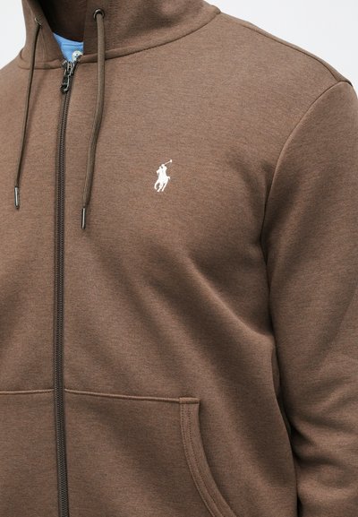 Smeđa zip-up dukserica napravljena od mekanog materijala. Ima kapuljaču, bočne džepove i bijeli logo na prsima. Tekstura, atletski kroj.