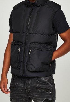 Czarna kamizelka puchowa z wieloma kieszeniami na zamki, noszona na czarnej koszulce z krótkim rękawem, w połączeniu z czarnymi przetartymi dżinsami z detalami na zamki.