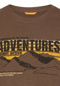 Brun T-shirt med teksten "ADVENTURES" over et gult og slidt sort bjergmotiv samt yderligere lille tekst med eventyrtema.