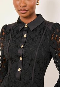 Mustast pitsist kleit, millel on ees nööbitav kuldsete nuppudega, kraega kaelus ja tekstuursete lillemustriga pikad varrukad.