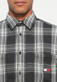 Juodai baltas langeliais marškinis, pagamintas iš tekstūruoto audinio, su mygtukais užsegamu apykakle, krūtinės kišene ir maža logotipo etikete kišenėje.