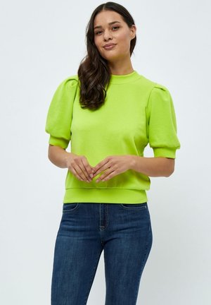 Hellgrüner Sweatshirt mit gepufften Kurzärmeln, geripptem Ausschnitt und Saum. Kombiniert mit dunkelblauen Jeans steht das Model mit gefalteten Händen.