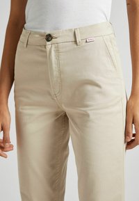 Pantaloni in misto cotone beige con una texture liscia, tasche laterali, chiusura con bottone e una piccola etichetta logo rosa vicino alla vita.