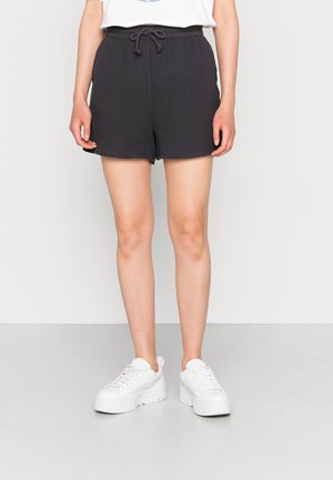 Persona vestida con pantalones cortos negros de cuerda ajustable y zapatillas de plataforma blancas, apoyada contra una pared y un suelo claros y lisos.