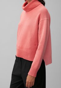 Vrouw in een roze coltrui met geribbelde manchetten en zoom, gecombineerd met een zwarte broek, tegen een effen grijze achtergrond.