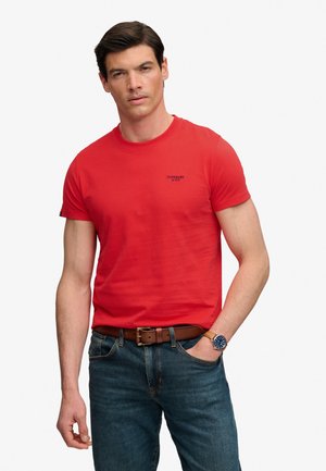 Mężczyzna w czerwonej koszulce Superdry, niebieskich dżinsach, brązowym pasku i zegarku na rękę, stojący z jedną ręką w kieszeni na białym tle.
