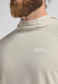 Chemise à capuche beige clair à manches longues avec une texture lisse. Présente un logo sur la poitrine et des coutures d'épaule sans couture pour le confort.