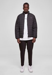 Black puffer jacket with a stand-up collar, featuring quilted texture, worn over a white t-shirt and paired with black trousers and white sneakers.
