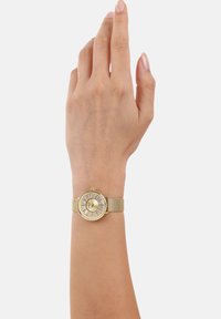 Goldene Uhr mit rundem Ziffernblatt, grobem Armband und strukturierten Stundenmarkierungen. Die Uhr hat ein minimalistisches Design und eine hervorstehende Krone für Einstellungen.