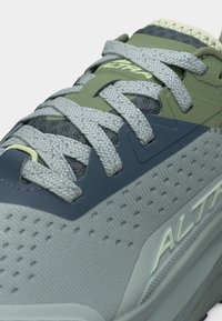 Chaussure de sport grise et verte avec des lacets gris clair et des détails perforés, arborant le nom de la marque "Altra" sur la languette et le côté.