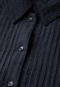 Camicia button-up in blu navy scuro realizzata in tessuto testurizzato con un aspetto stropicciato. Presenta un colletto aperto e piccoli bottoni neri.