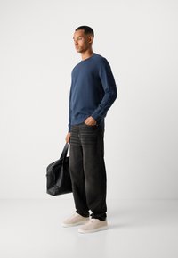 Mann iført en marineblå hettegenser, vide svarte jeans, beige sneakers, holder en svart lærveske og står mot en ensfarget hvit bakgrunn.