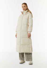 Langer, beiger Daunenmantel mit Stehkragen, durchgehendem Reißverschluss und seitlichen Knöpfen. Verfügt über eine gesteppte Textur und einen übergroßen Schnitt.