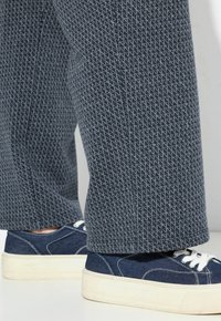 Blå texturerade byxor tillsammans med mörkblå canvas sneakers med vita snören och tjocka vita sulor på en neutral bakgrund.