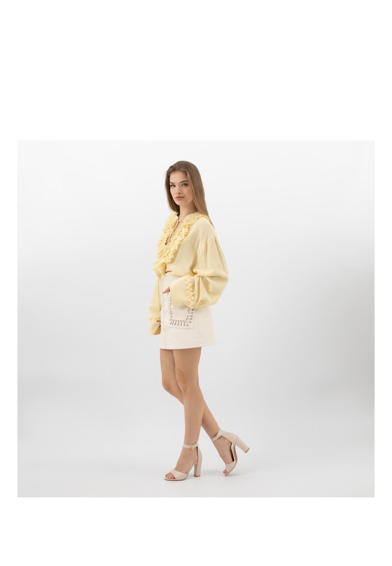 Jonge vrouw die een lichtgele blouse met ruches draagt, een beige rok met een gestikte zak en beige sandalen met hoge hakken, staand in zijaanzicht.