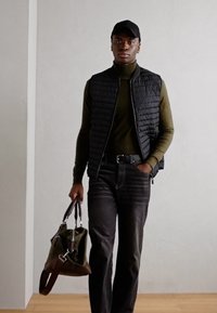 Zwart gewatteerd vest over een donkergroen coltrui, gecombineerd met donkere jeans. Houdt een donkergroene tas met bruine accenten vast en draagt een zwarte pet.