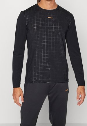 Man draagt een zwart, langärmelig shirt met ruitpatroon en oranje "BOSS"-logo, gecombineerd met een zwarte broek met eveneens een oranje "BOSS"-logo.