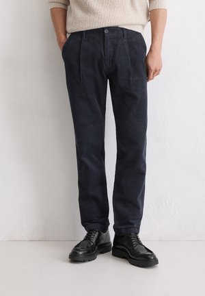 Marine blauwe corduroy broek met voorplooien, met een recht gesneden ontwerp en een knoopsluiting. Draagt met zwarte veterschoenen.