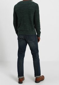 Dunkelgrüner, strukturierter Pullover mit Rundhalsausschnitt, kombiniert mit dunkelblauen Jeans und braunen Schuhen, von hinten betrachtet.