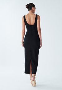 Black fitted dress with a low back, knee-length slit, and wide straps. Smooth texture, featuring a central zipper at the back.