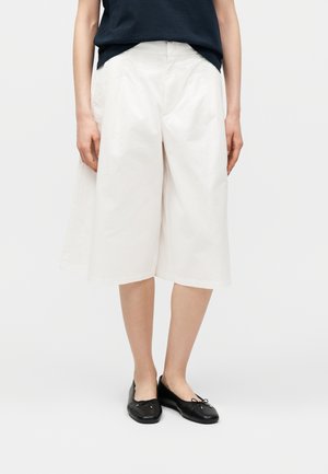 Kvinde iført løse hvide knælange shorts, sorte balletsko og en sort skjorte stoppet ind, stående mod en ensfarvet baggrund.