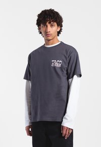 Grå bomullströja med korta ärmar, med en tryckt rosa grafik på bröstet; lager över en långärmad vit skjorta.