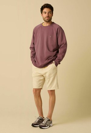 Homme debout, mains dans les poches, portant un sweat mauve, un short beige et des chaussures de running grises sur fond beige.
