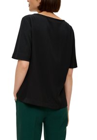 Zwarte top met korte mouwen en een ronde halslijn, met een scalloped afwerking aan de achterkant en een losse pasvorm. Gepaard met groene broek.