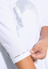 Weißes T-Shirt mit langen Ärmeln, das auf der Vorderseite ein Paillettenmuster und einen dekorativen Paillettenbesatz an den Ärmelabschlüssen aufweist.